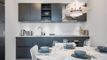 Una cocina moderna con armarios oscuros, una mesa de comedor blanca con cuencos azules y copas de vino, y una lámpara colgante arriba.