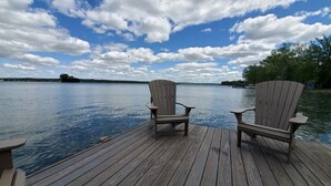 A wooden deck with two Adirondack chairs facing a calm lake. - 1818 Modern Farm House with Pool, Hot Tub and a large permanent dock on the lake (Canandaigua)
