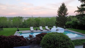 A pool with lounge chairs and a slide, surrounded by greenery and a view of a distant cityscape. - 1818 Modern Farm House with Pool, Hot Tub and a large permanent dock on the lake (Canandaigua)