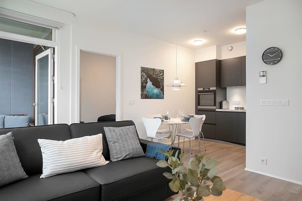 Una sala de estar moderna con un sofá negro, una zona de comedor con mesa y sillas blancas, y una cocina con armarios oscuros y un reloj en la pared.