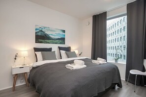 Moderni hotellihuone, jossa on suuri sänky, yöpöytä lampun kanssa, ikkunasta avautuva näkymä kaupunkimaisemaan ja taulu seinällä.