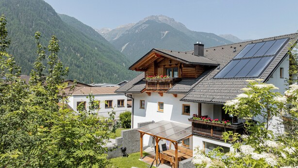 A chalet with a balcony, solar panels, and a mountain backdrop. - Landhaus Waldhart - Cozy apartment with garden & Summer Card (Umhausen)