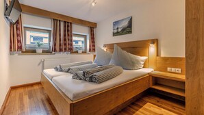 A wooden bed with checkered bedding, a bedside table with a lamp, a window with curtains, and a painting on the wall. - Landhaus Waldhart - Cozy apartment with garden & Summer Card (Umhausen)
