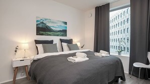 Una habitación de hotel moderna con una cama grande, una mesita de noche con lámpara, una ventana con cortinas y vistas a la ciudad.