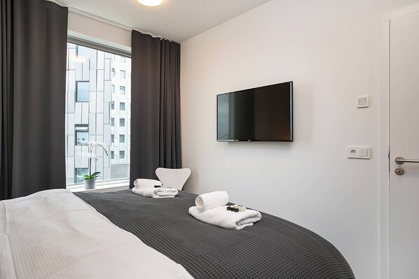 A hotel room with a bed, a TV mounted on the wall, a window with curtains, and a view of buildings outside. - SIF Apartments - 2 bedrooms with 2 bathrooms (Reykjavík)