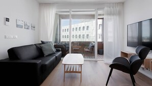 Una sala de estar moderna con un sofá de cuero negro, una mesa de centro blanca y sillas negras. Hay un televisor de pantalla plana grande sobre un soporte de madera. La habitación tiene ventanas amplias con cortinas blancas, que ofrecen vistas de un edificio exterior.