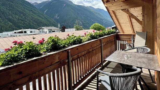 A wooden balcony with a view of mountains, a table, and chairs.
