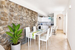 Une cuisine moderne avec des armoires blanches, un mur d’accent en pierre et un coin repas comprenant des chaises blanches et une table en verre.