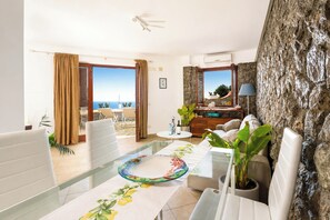 Una sala de estar moderna con una mesa de vidrio, sofás blancos, vista al mar y una pared de piedra.