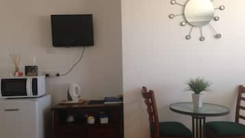 A compact kitchen with a microwave, kettle, and small cabinet. A TV is mounted on the wall, and a round mirror with a sunburst design hangs above a dining table with two chairs.