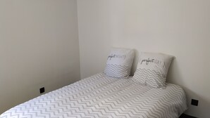 Een net opgemaakt bed met een sprei met een zigzaggemotief, twee kussens met tekst, en een wandlamp.