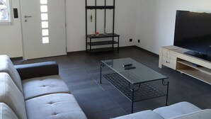 Een moderne woonkamer met een hoekbank, een glazen salontafel, een zwart metalen plankunit en een televisie op een houten standaard.