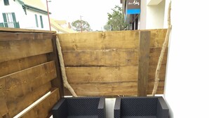 Een houten terras met een ingebouwde zwarte zithoek met kussens en een kleine tafel met een glas en een plant.