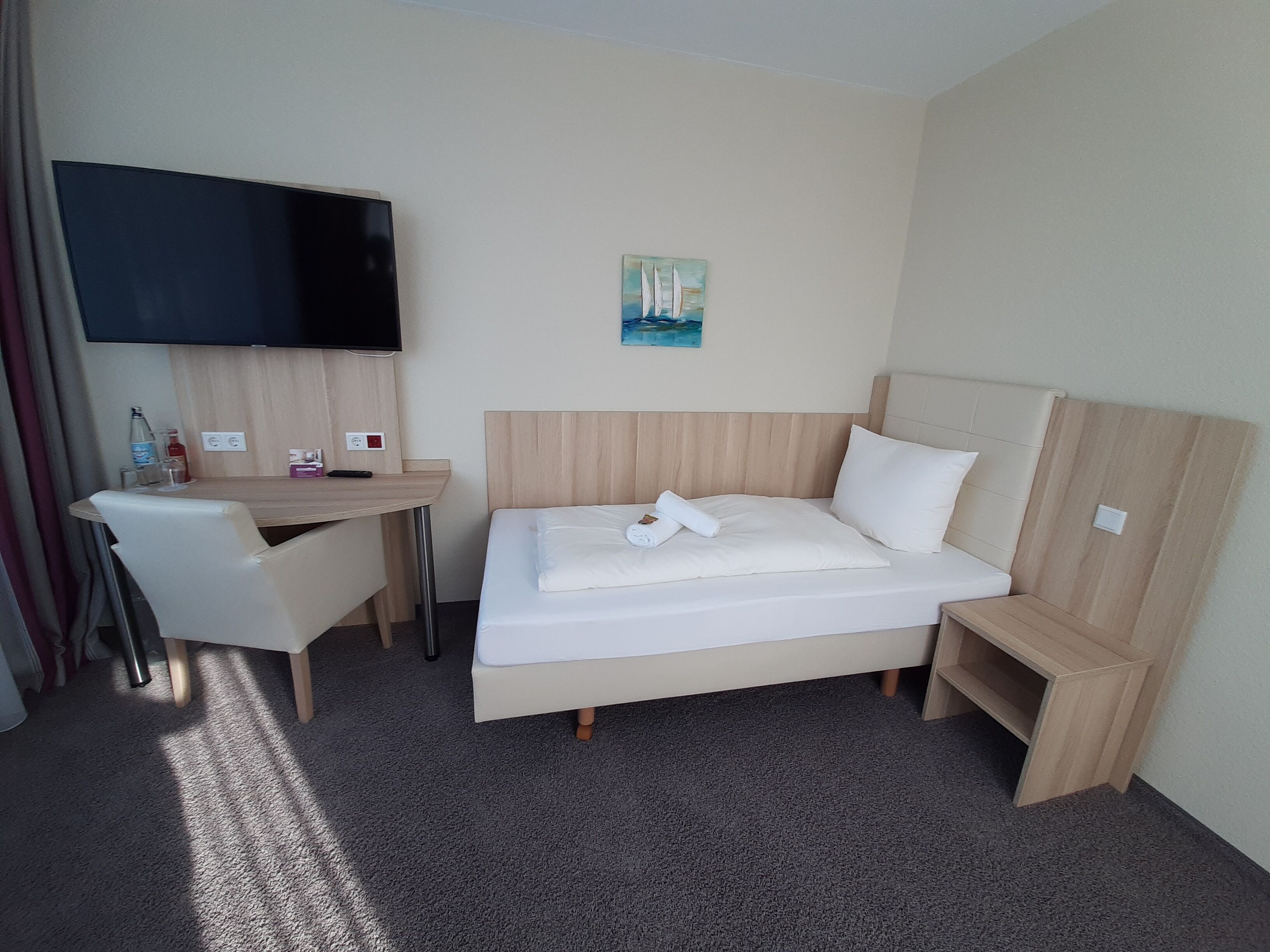 A hotel room with a bed, a desk, a chair, a TV, and a painting on the wall.