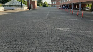 Un área de estacionamiento con superficie pavimentada, un pequeño edificio con techo de vidrio y una fila de coches aparcados.