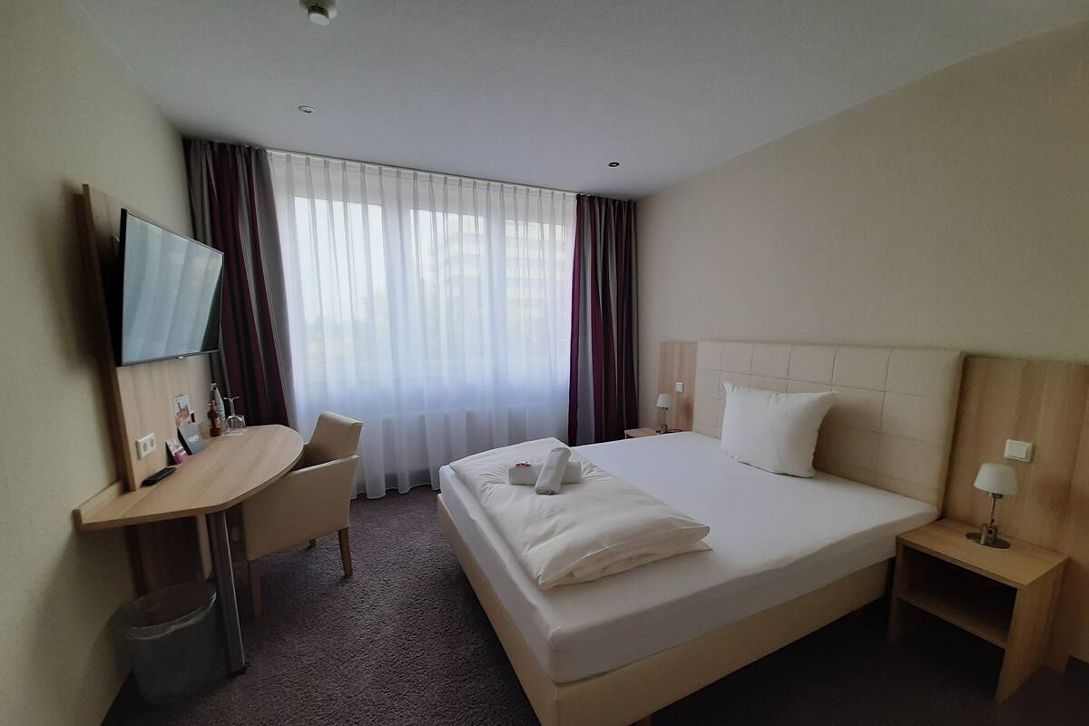 A hotel room with a bed, a desk, a chair, a TV, and a window with curtains.