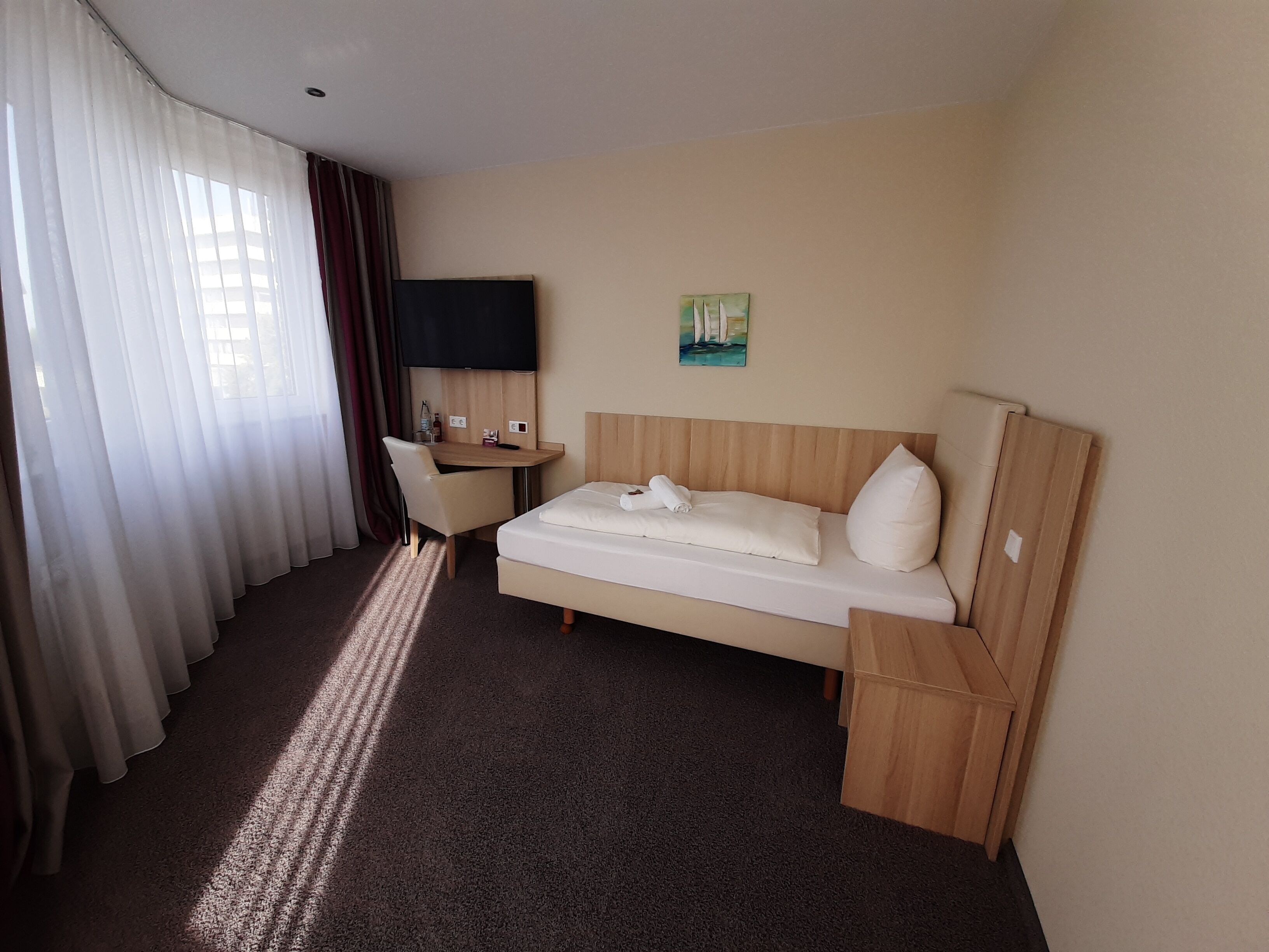A hotel room with a bed, a desk, a chair, a TV, and a painting on the wall.