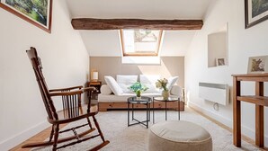 A cozy room with a wooden rocking chair, a sofa with white cushions, a small table with a plant, and a large window providing natural light.