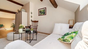A cozy, well-lit room with a sloped ceiling, a white sofa with a green leaf pillow, a wooden chair, a small round table with a vase of flowers, and a framed picture on the wall.
