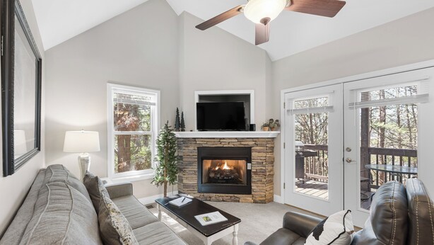 A living room with a stone fireplace, a ceiling fan, and a large window. - Lark’s Nest Mtn View - See Yonah Mountain from the Front Porch! (Helen)