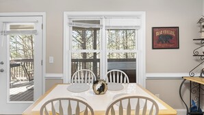 A dining area with a white table and chairs, a framed bear crossing sign on the wall, and a view of trees through the windows. - Lark’s Nest Mtn View - See Yonah Mountain from the Front Porch! (Helen)