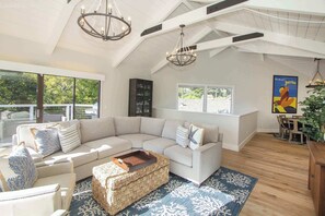 A spacious living room with a large sectional sofa, a coffee table, and a chandelier. There are large windows providing natural light and a view of the outdoors.