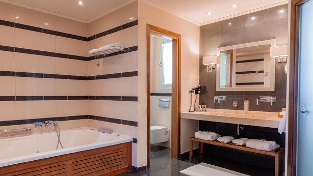 A modern bathroom with a large bathtub, a vanity with a mirror, and a bench with towels. - Restia (Corfu)