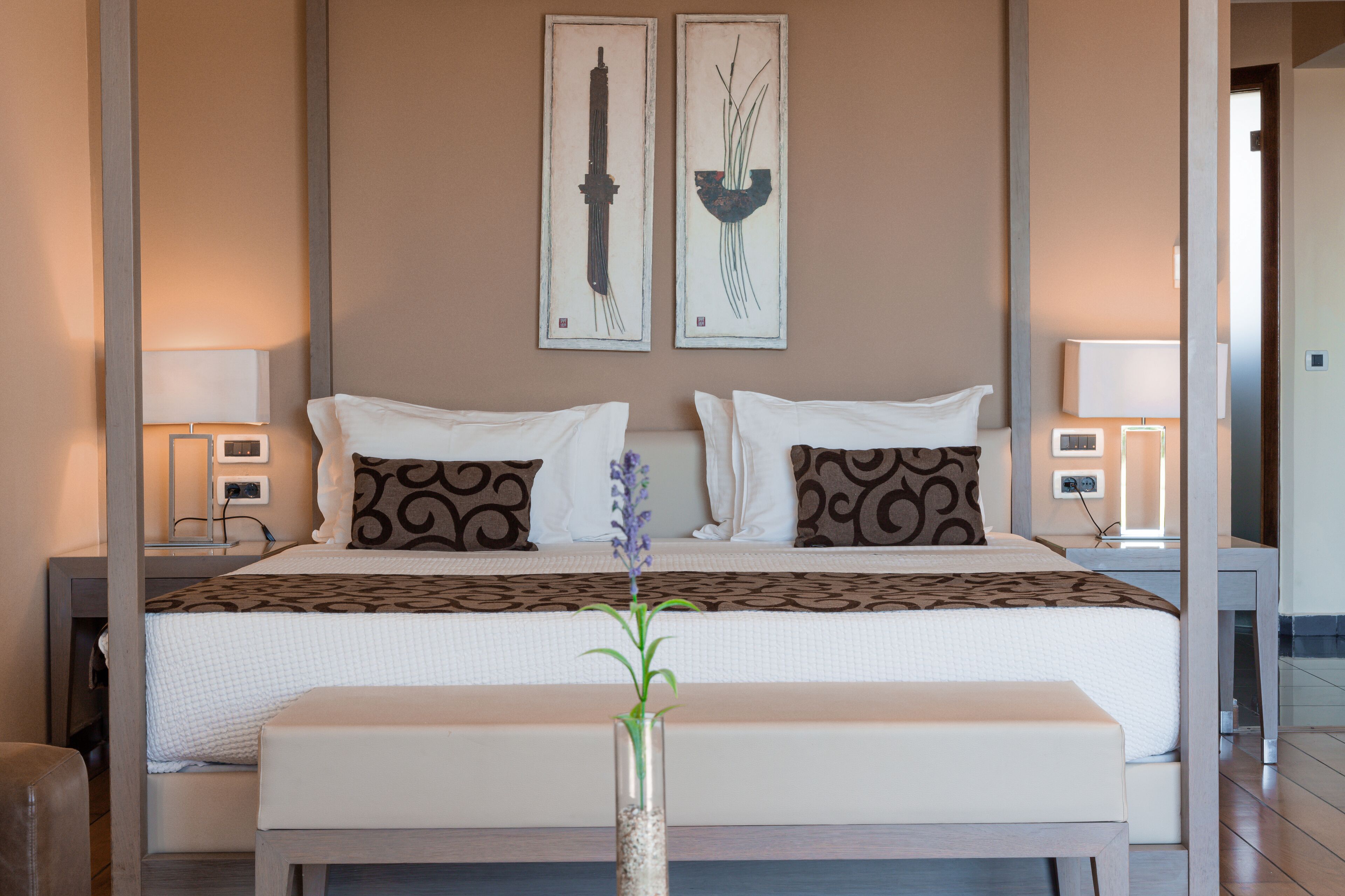 A modern hotel room with a large bed, two bedside tables with lamps, a vase with a plant, and artwork on the walls.