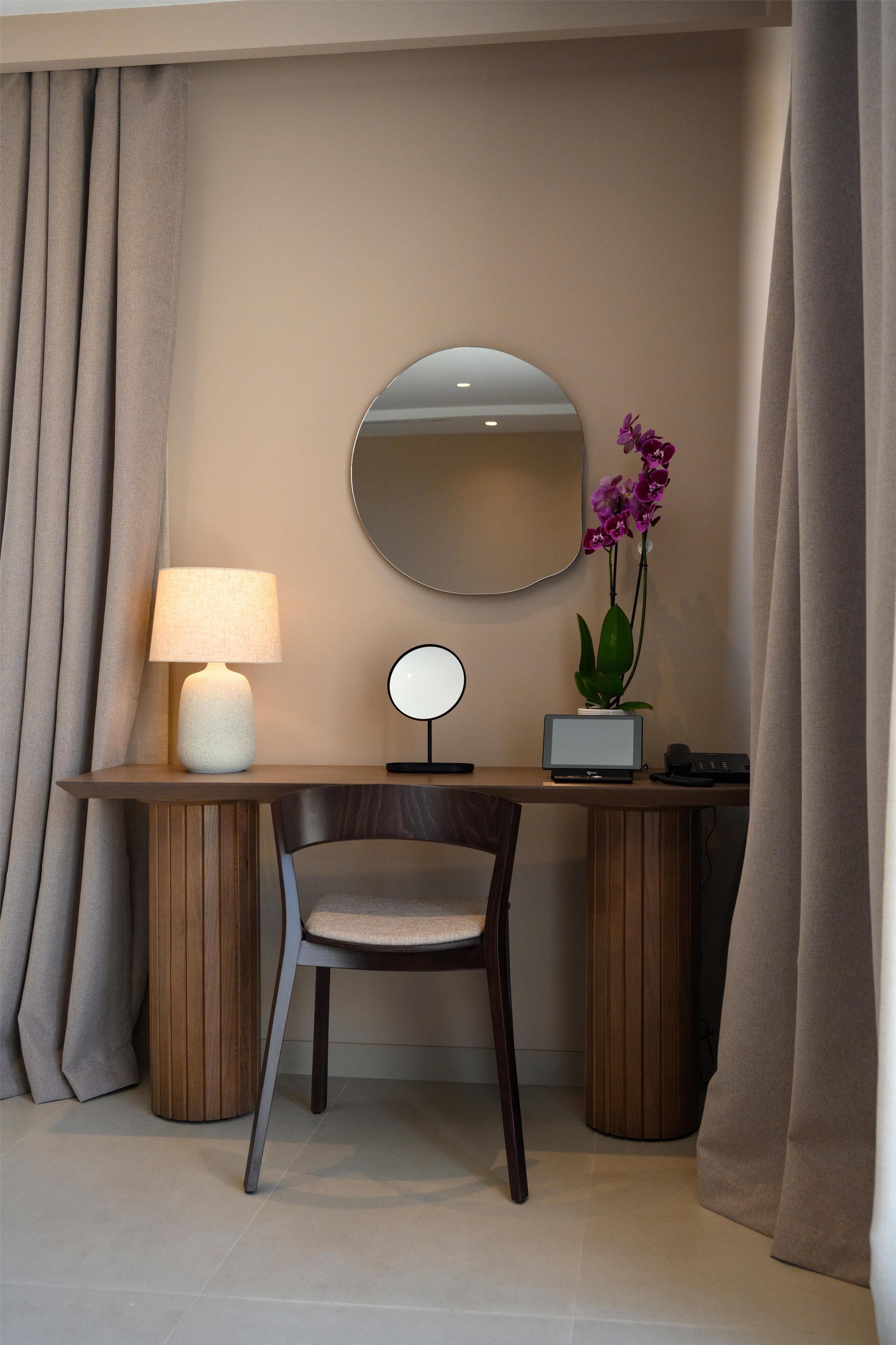 A hotel room with a desk, chair, lamp, mirror, and orchid plant.