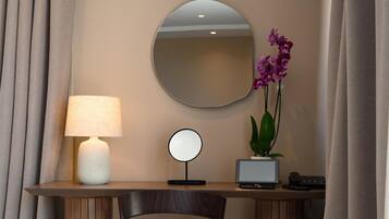 A hotel room with a desk, chair, lamp, mirror, and orchid plant.