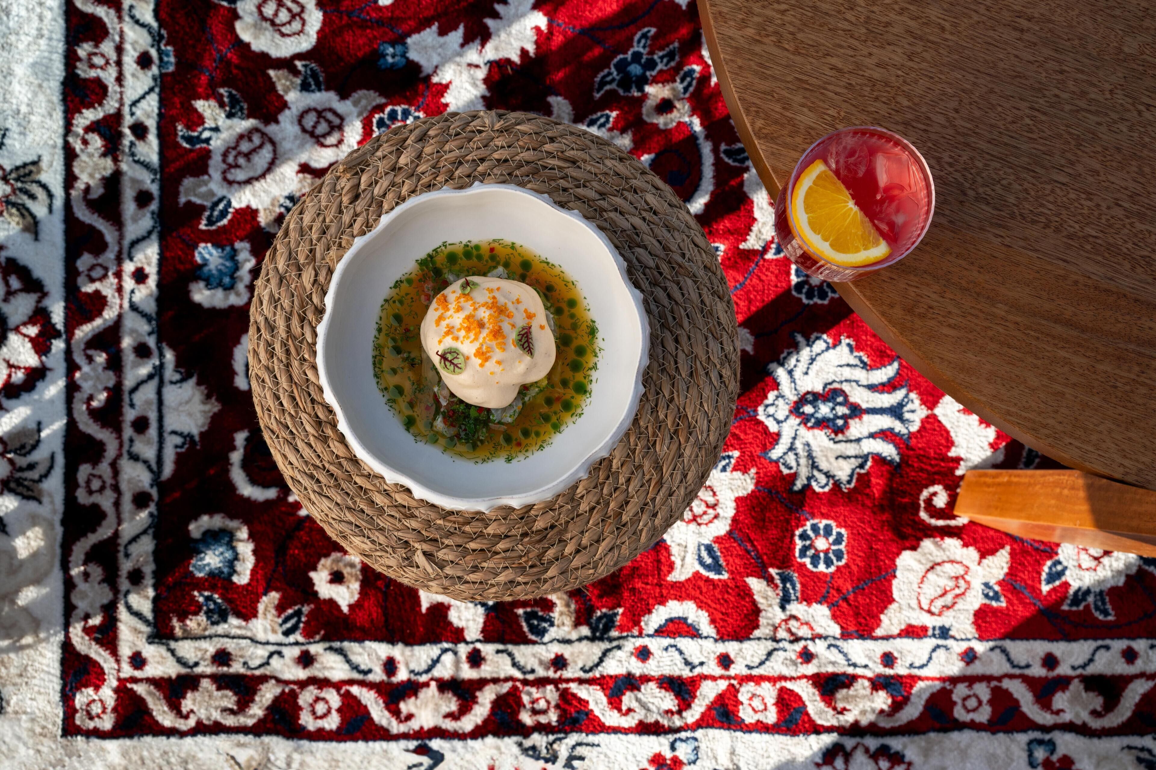 Una estera tejida con un plato de comida y un vaso de bebida sobre una alfombra estampada.