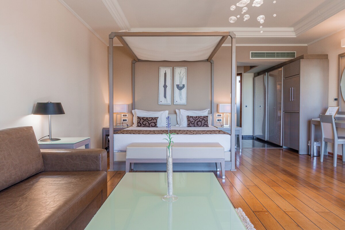 A bedroom with a four-poster bed, bedside tables, a nightstand, a sofa, and a dining area with a chair.