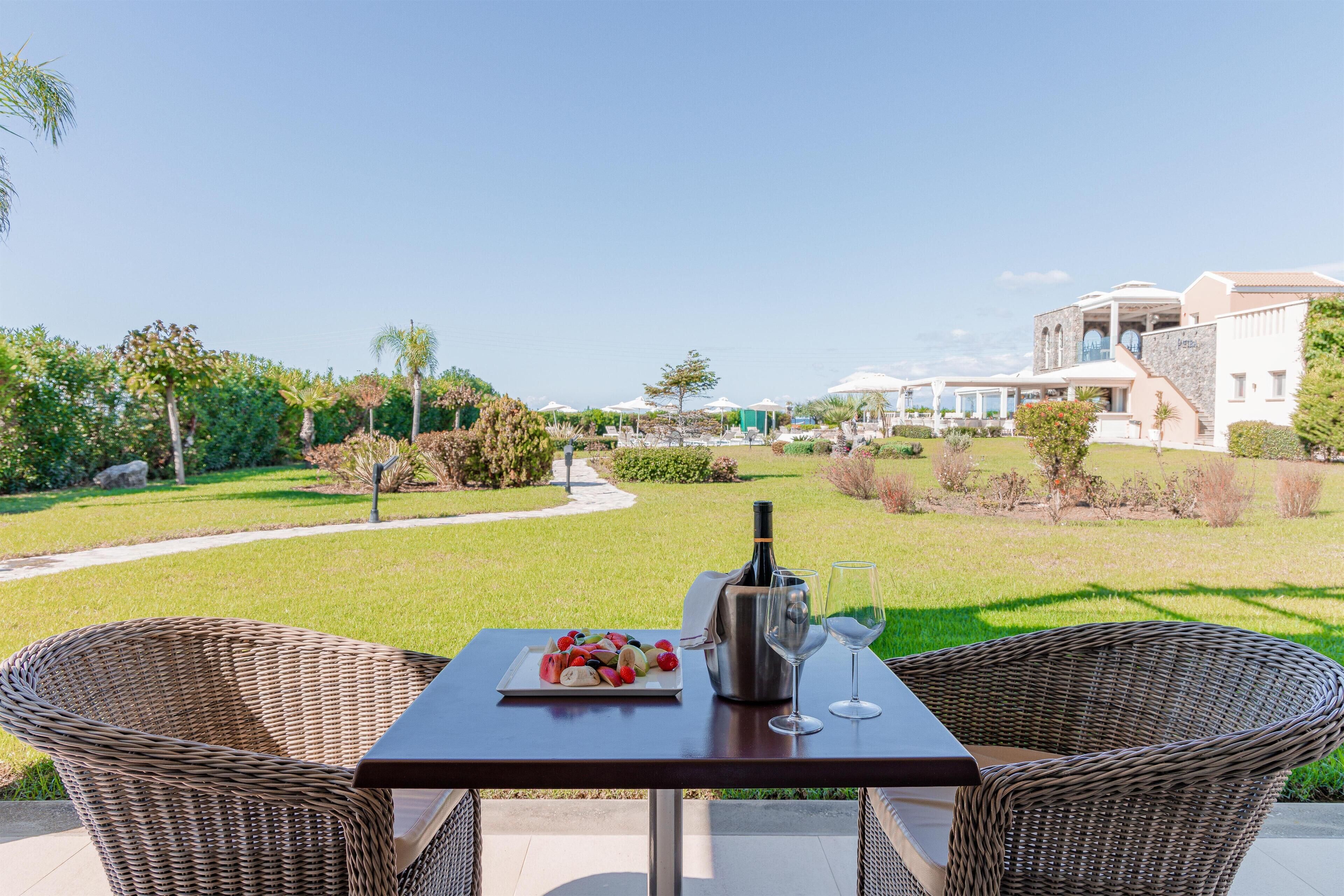 Una mesa de comedor con sillas de mimbre, una botella de vino y una copa en un patio con vistas a un césped cuidado y una piscina junto a un edificio.