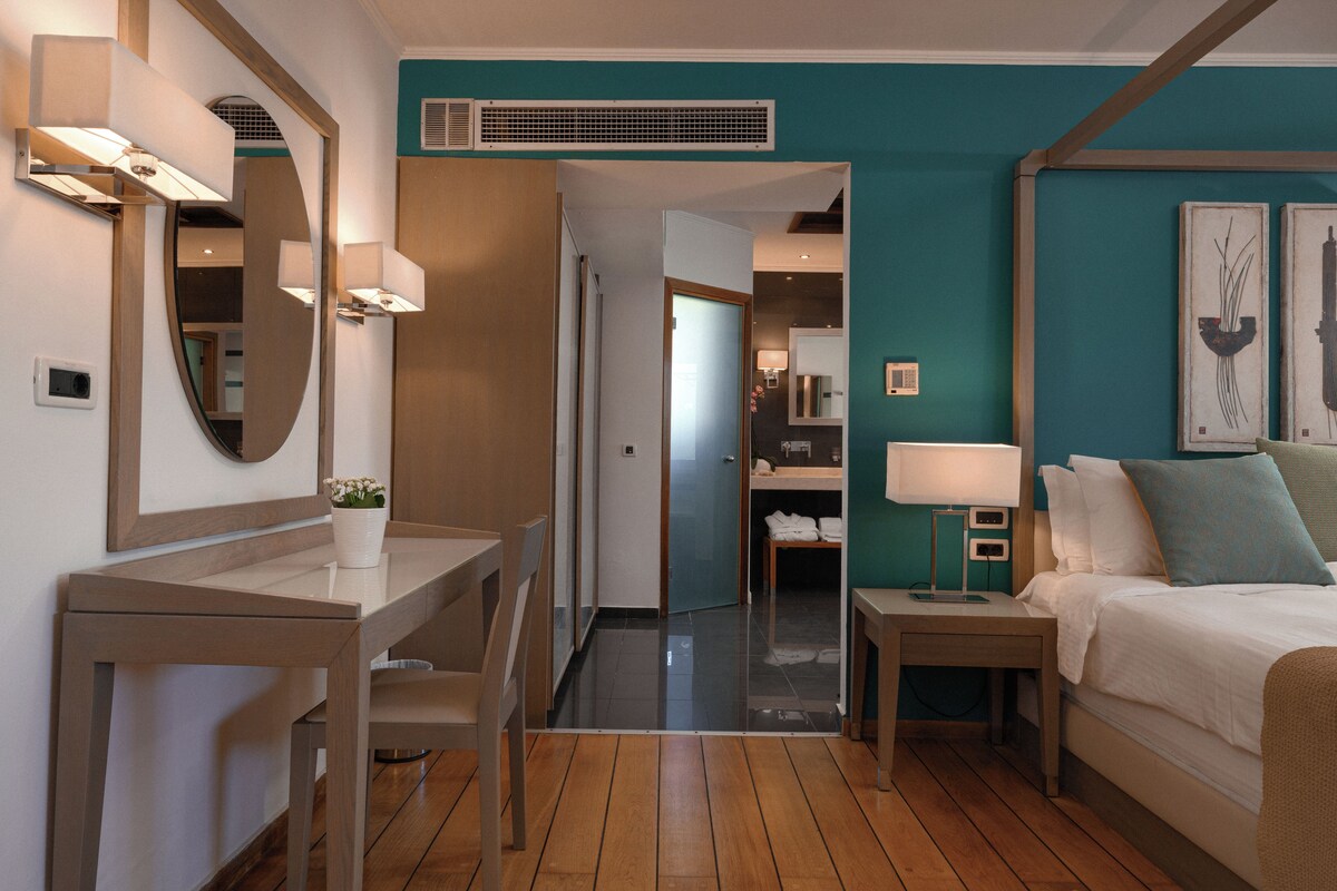 A hotel room with a bed, bedside table, lamp, mirror, and a small table with a potted plant.