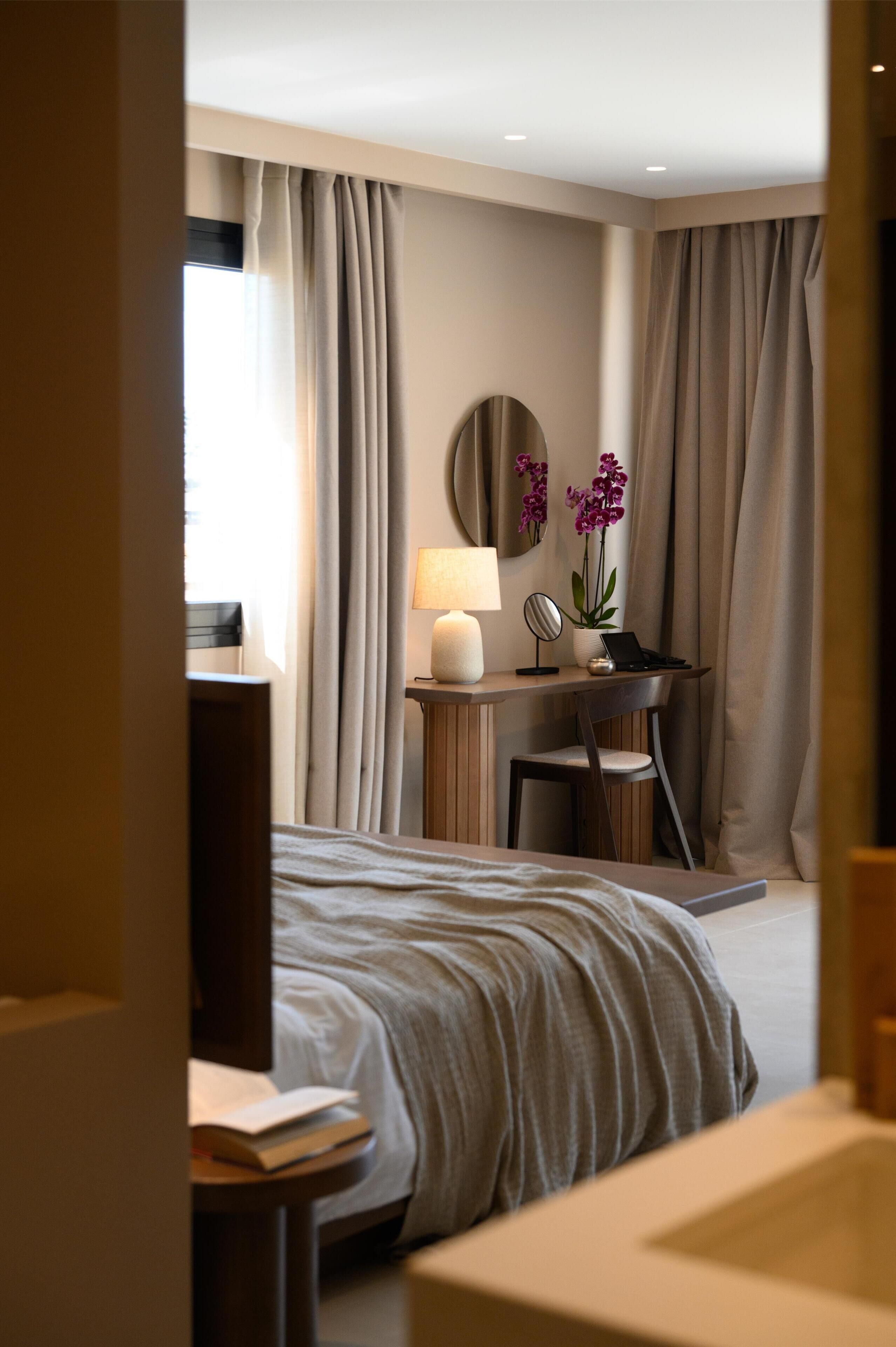 Ein Hotelzimmer mit einem Bett, einem Schreibtisch mit einem Laptop, einer Lampe, einem Spiegel und einer Topforchidee.