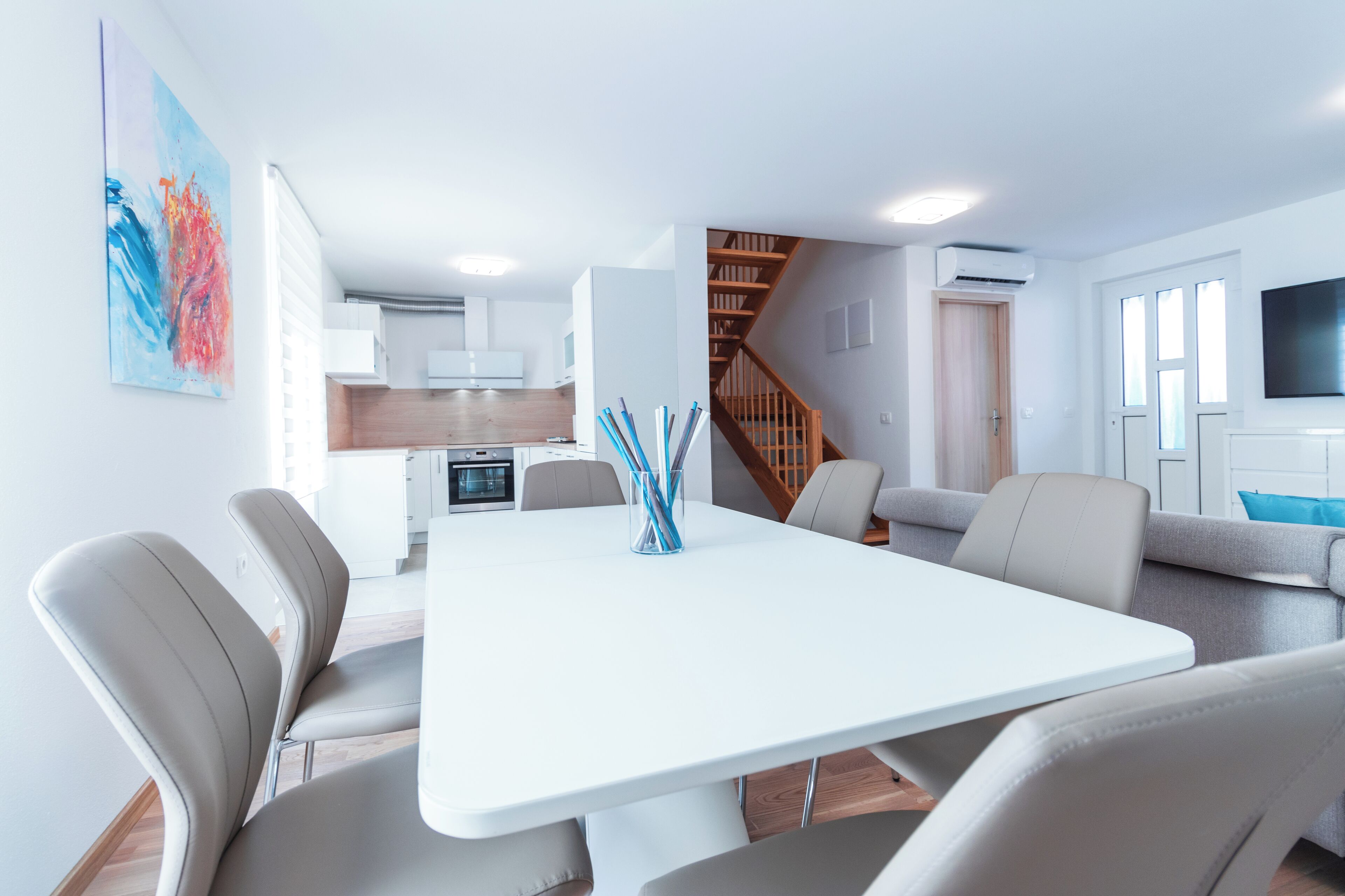 A modern living room with a white dining table, grey sofas, and a staircase leading to an upper floor.