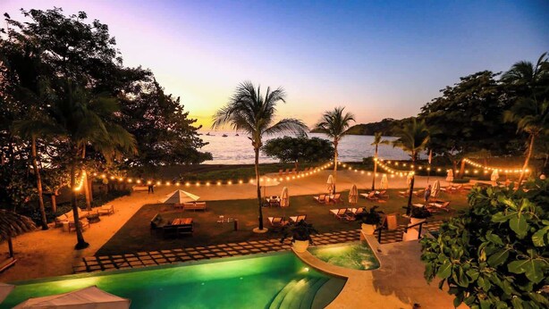 A pool area with lounge chairs and umbrellas, surrounded by trees and string lights. - Chic Minimalist Condo with Swimming Pool, Hot Tub, and Beach Club Access (Playa del Coco)