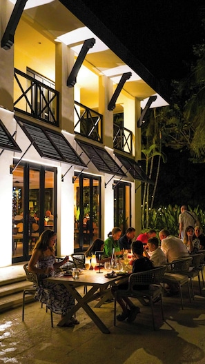 An outdoor dining area with tables and chairs, a building with a balcony, and a tropical garden in the background. - Picturesque Condo with Spectacular Ocean Views & Beach Club Access (Playa del Coco)
