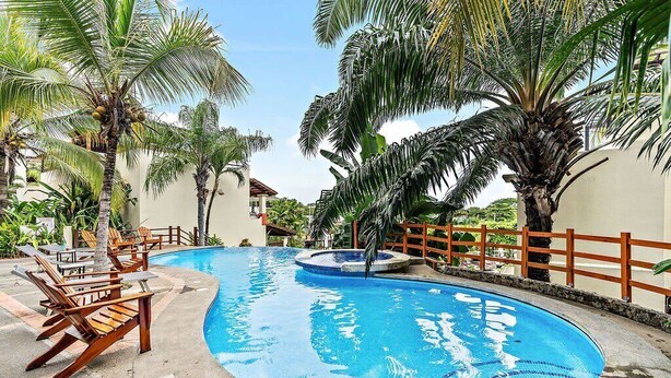 A swimming pool surrounded by palm trees and wooden chairs. - Colorful & Private 2-Bedroom Condo Just a Short Walk from the Beach (Coco)
