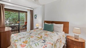 A hotel room with a bed, a nightstand, a ceiling fan, a window with curtains, and a balcony with chairs. - Colorful & Private 2-Bedroom Condo Just a Short Walk from the Beach (Coco)