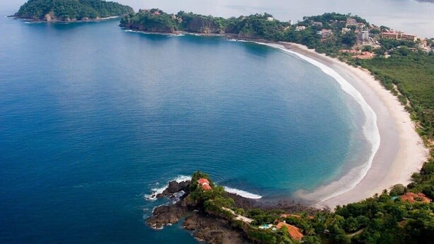 An aerial view of a coastal area with a curved beach, rocky outcrops, and scattered buildings. - Chic Cliffside Condo w/ Two Balconies Offering Spectacular Ocean & Mountain Views (Playa Flamingo)