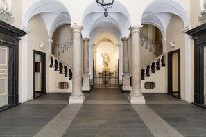 A grand hallway with a central statue, flanked by columns and a staircase leading to an upper level.