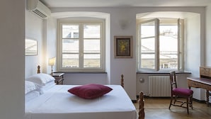 Ein Schlafzimmer mit einem Bett, einem Sessel, einem Schreibtisch und zwei Fenstern.