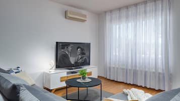 Un soggiorno moderno con divano, tavolino, televisore e un'ampia finestra con tende leggere.