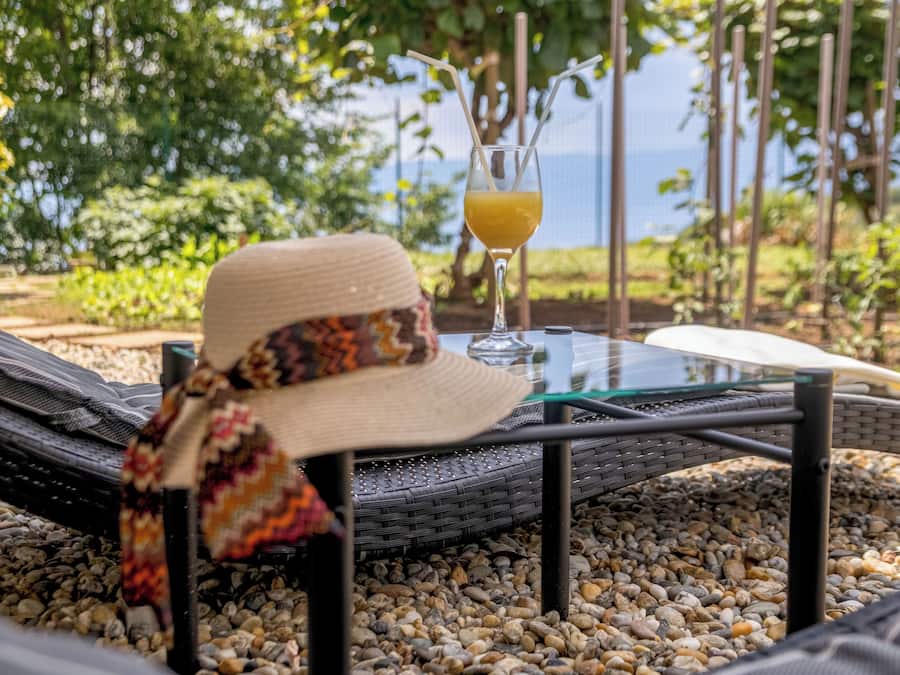 A table with a glass of drink, a hat, and a woven chair in a garden setting.