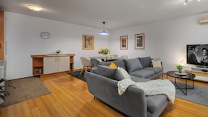A modern living room with a grey sofa, wooden flooring, a dining table with chairs, a small kitchen area, and a flat-screen TV. - Terra Magica Deluxe Apartment OR Room with Private Parking, Terrace and Sea View (Cavle)