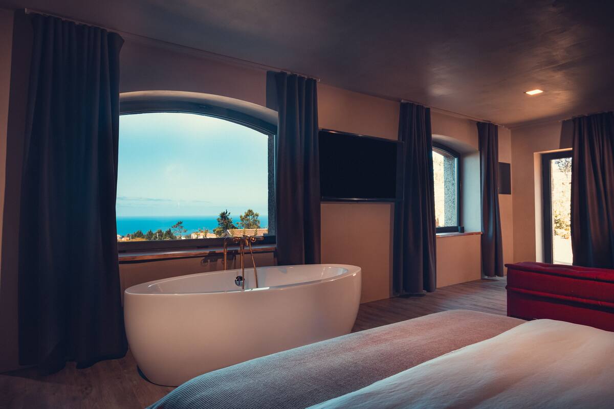 A modern hotel room with a large bathtub, a bed, a TV mounted on the wall, and a view of the sea.