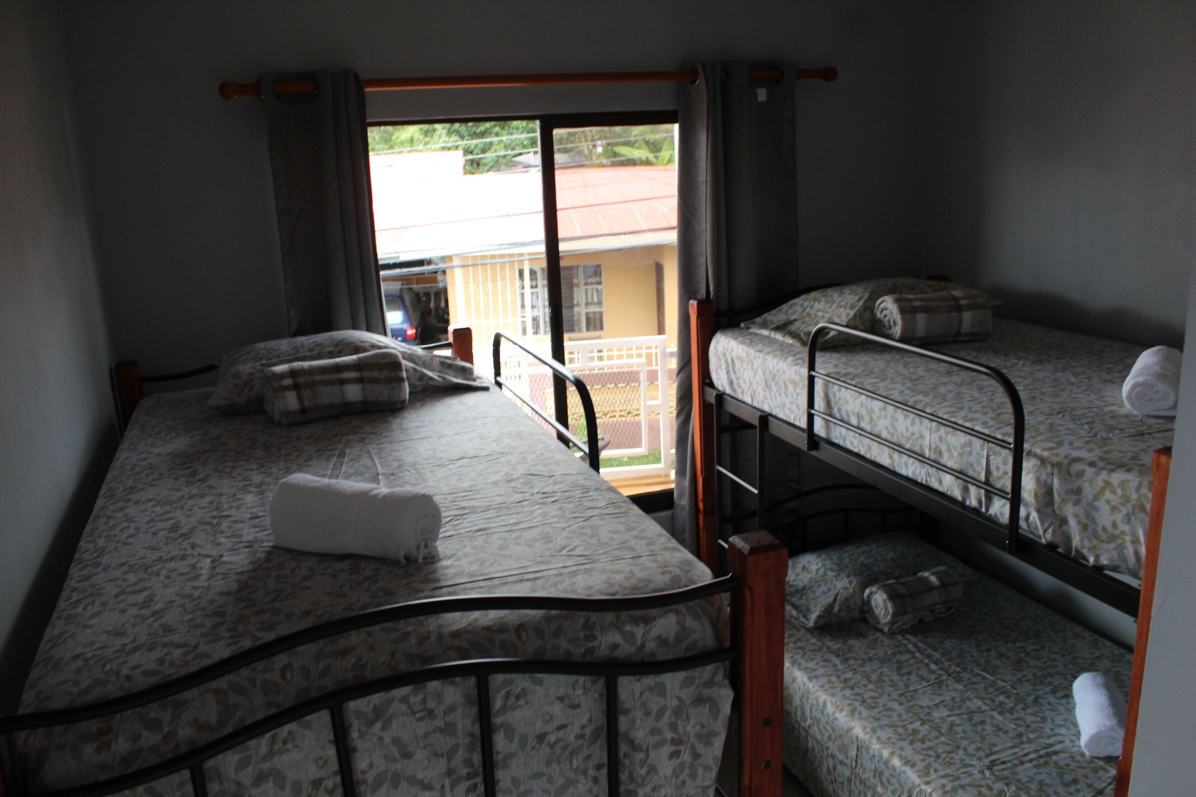 A dormitory room with two bunk beds, a window with curtains, and a view of a building outside.