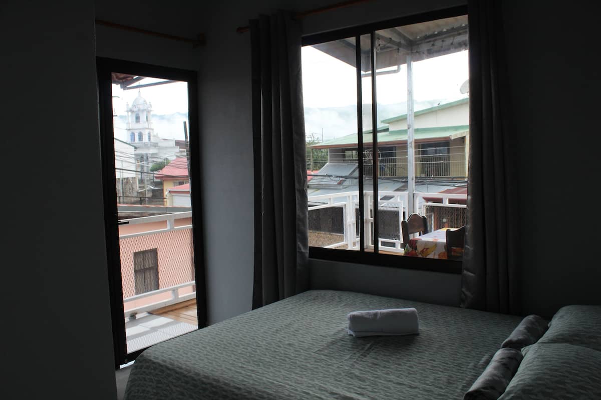 A hotel room with a bed, a window with curtains, and a view of buildings outside.