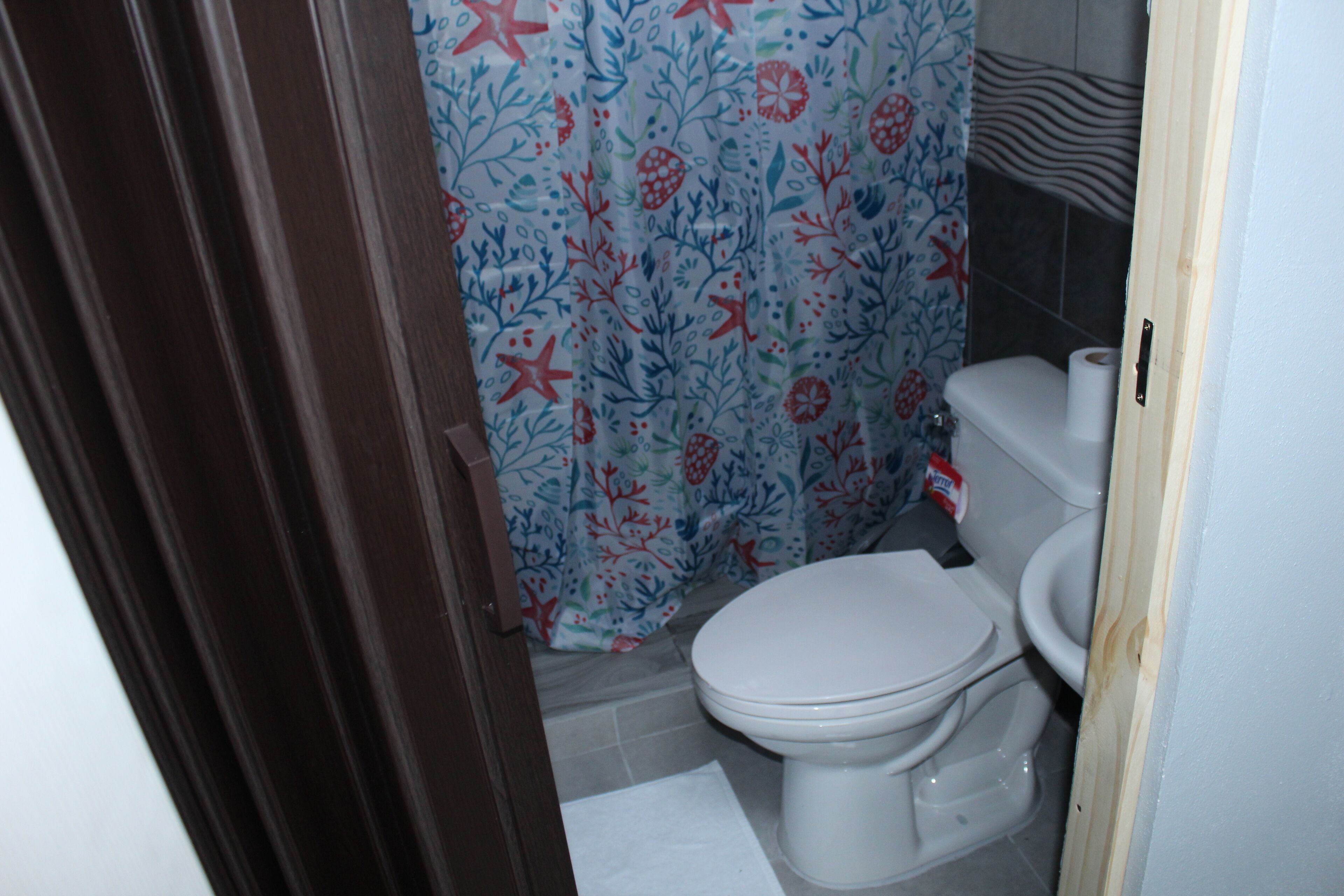 A bathroom with a toilet, a shower curtain with a floral pattern, and a white mat on the floor.